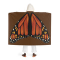 Model standing with back facing camera wearing brown hooded sherpa blanket showing monarch butterfly wings printed across the back and cream sherpa‑lined hood.