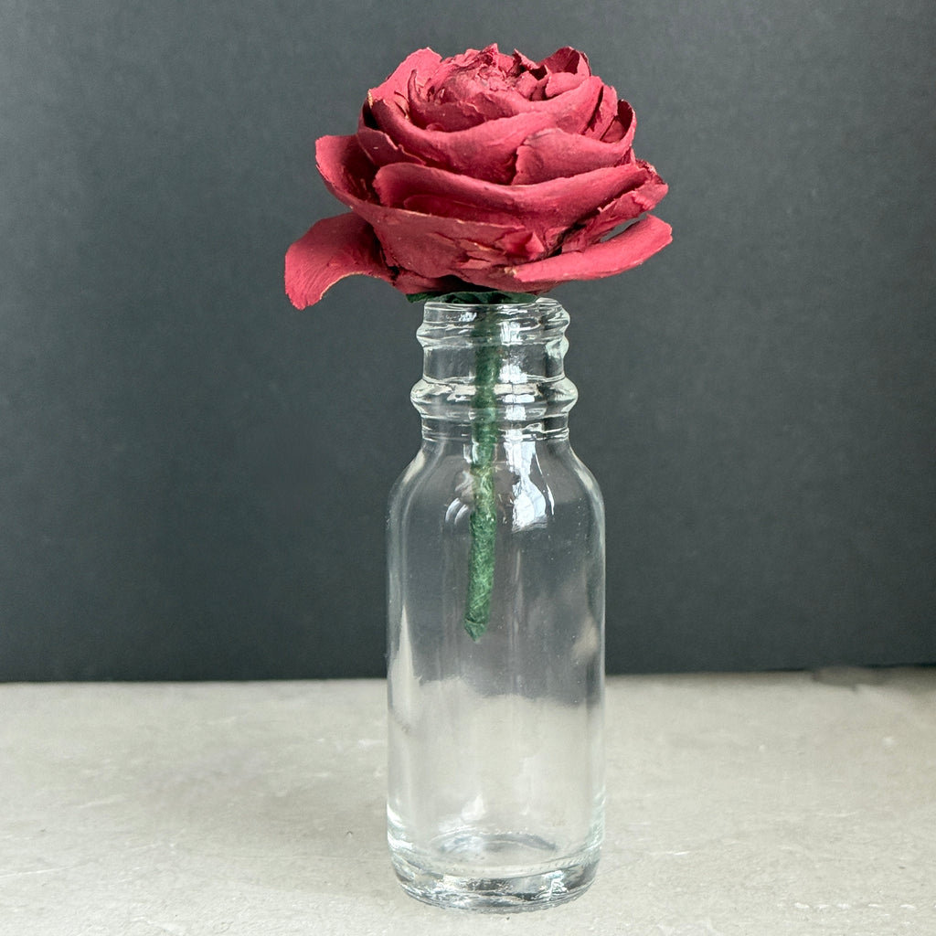 Deep burgundy Cedar Rose in a small clear glass bottle against a dark gray background.